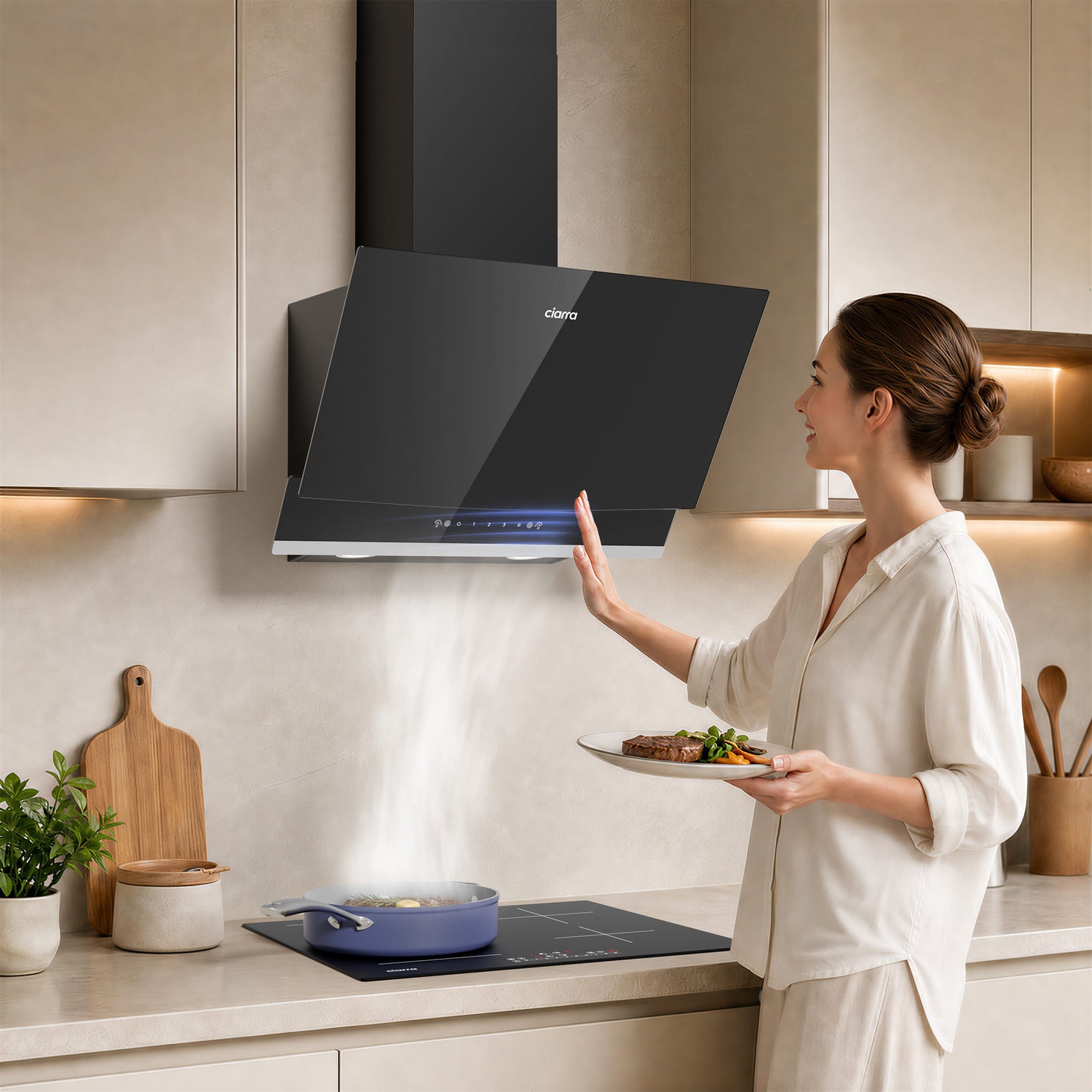 A woman in a white shirt stands in a modern kitchen holding a plate of food, reaching toward a black glass CIARRA cooker hood mounted above a cooktop. The kitchen has light beige cabinets, a countertop with a cutting board, plant, and jars, and warm under-cabinet lighting.