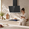 A woman in a bright kitchen stands beside a black induction cooktop and a black CIARRA cooker hood mounted on a beige wall. The scene is softly lit with natural shadows, and kitchen items like a cutting board, a vase of flowers, and a bowl of salad are visible.