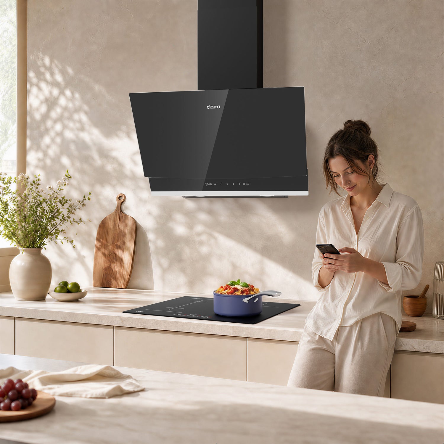 A woman in a bright kitchen stands beside a black induction cooktop and a black CIARRA cooker hood mounted on a beige wall. The scene is softly lit with natural shadows, and kitchen items like a cutting board, a vase of flowers, and a bowl of salad are visible.