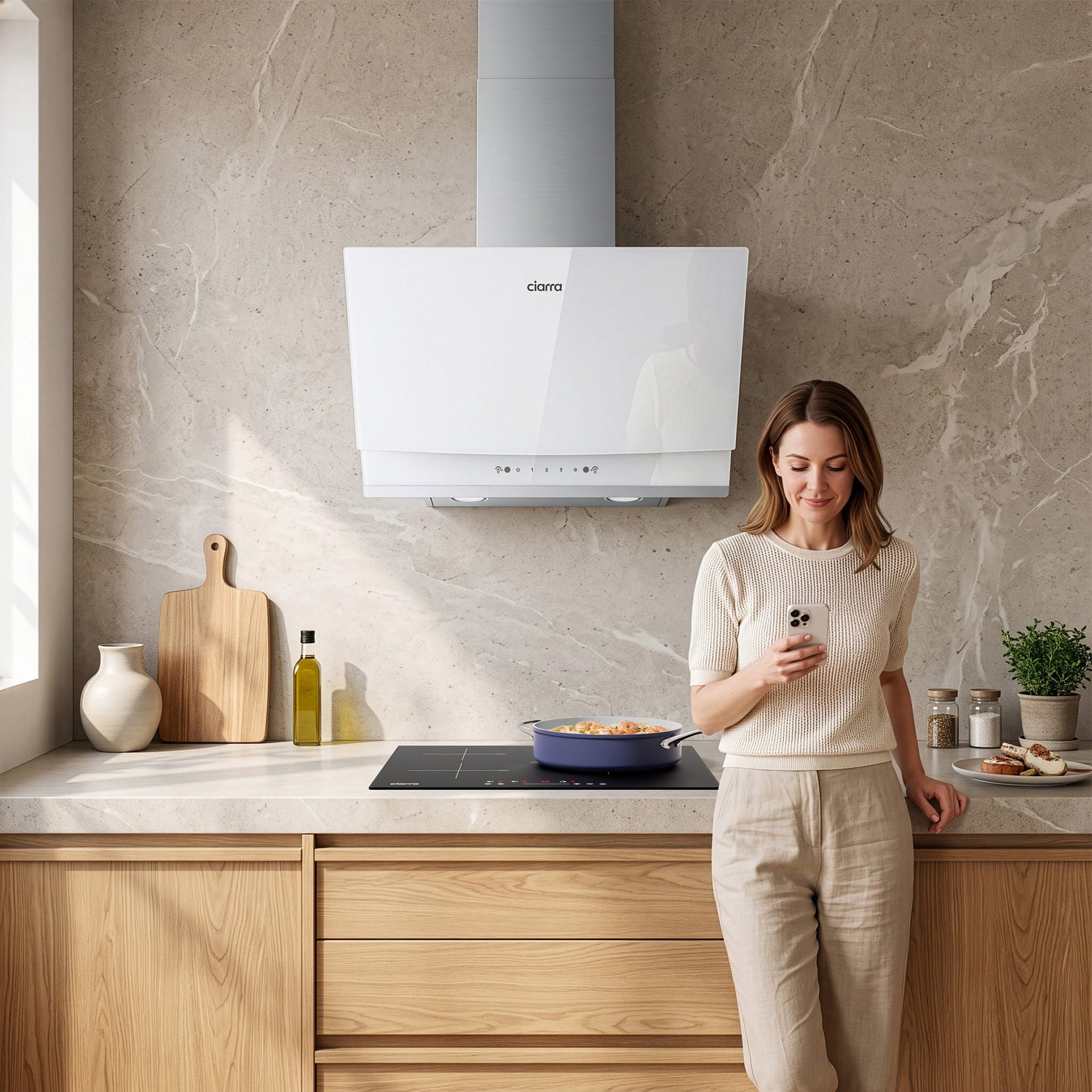 A woman stands in a modern kitchen next to a white CIARRA cooker hood mounted on a stone wall above a black induction cooktop.